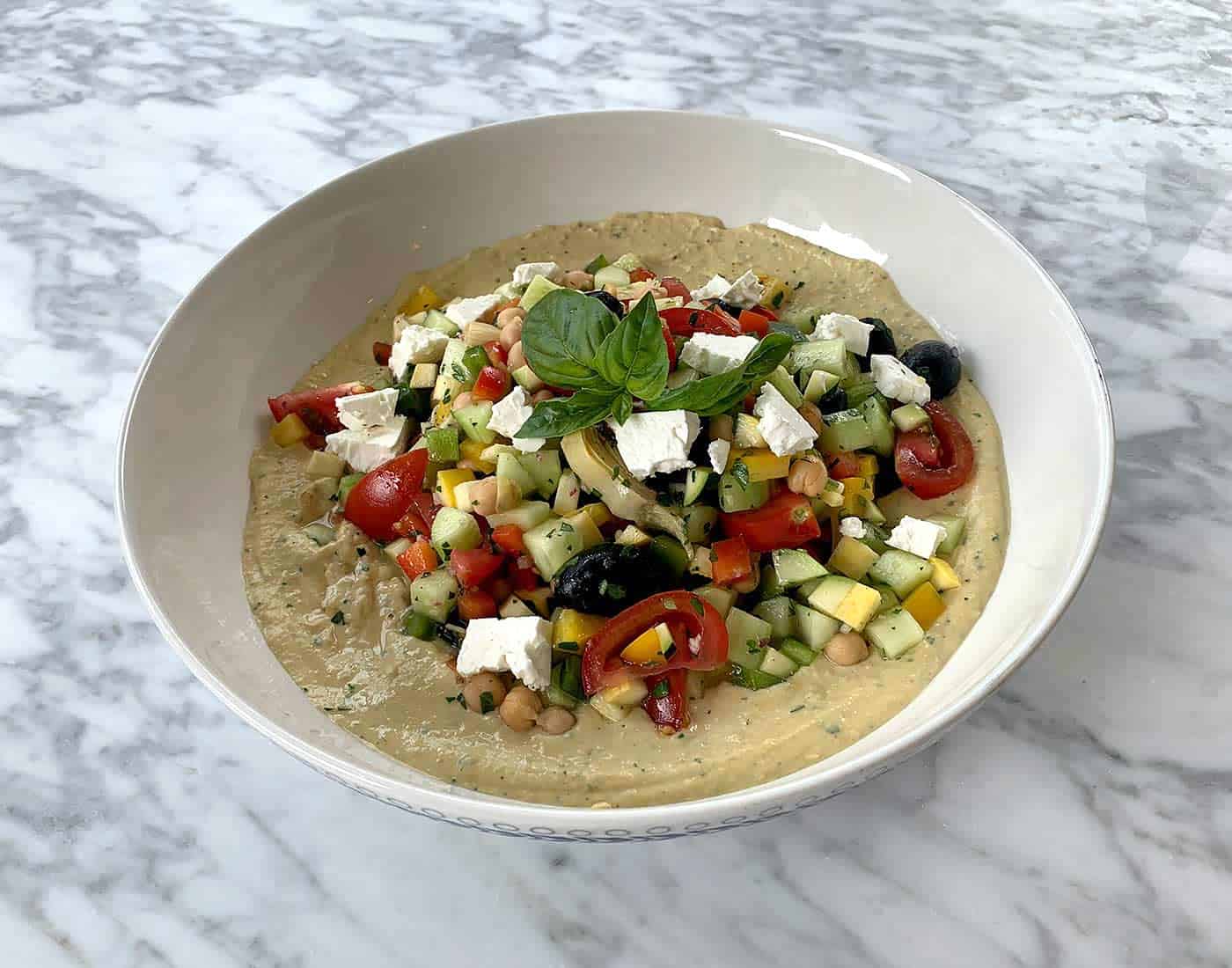 Hummus topped with chickpeas, cucumber, yellow bell peppers, tomatoes, feta, basil, and olives