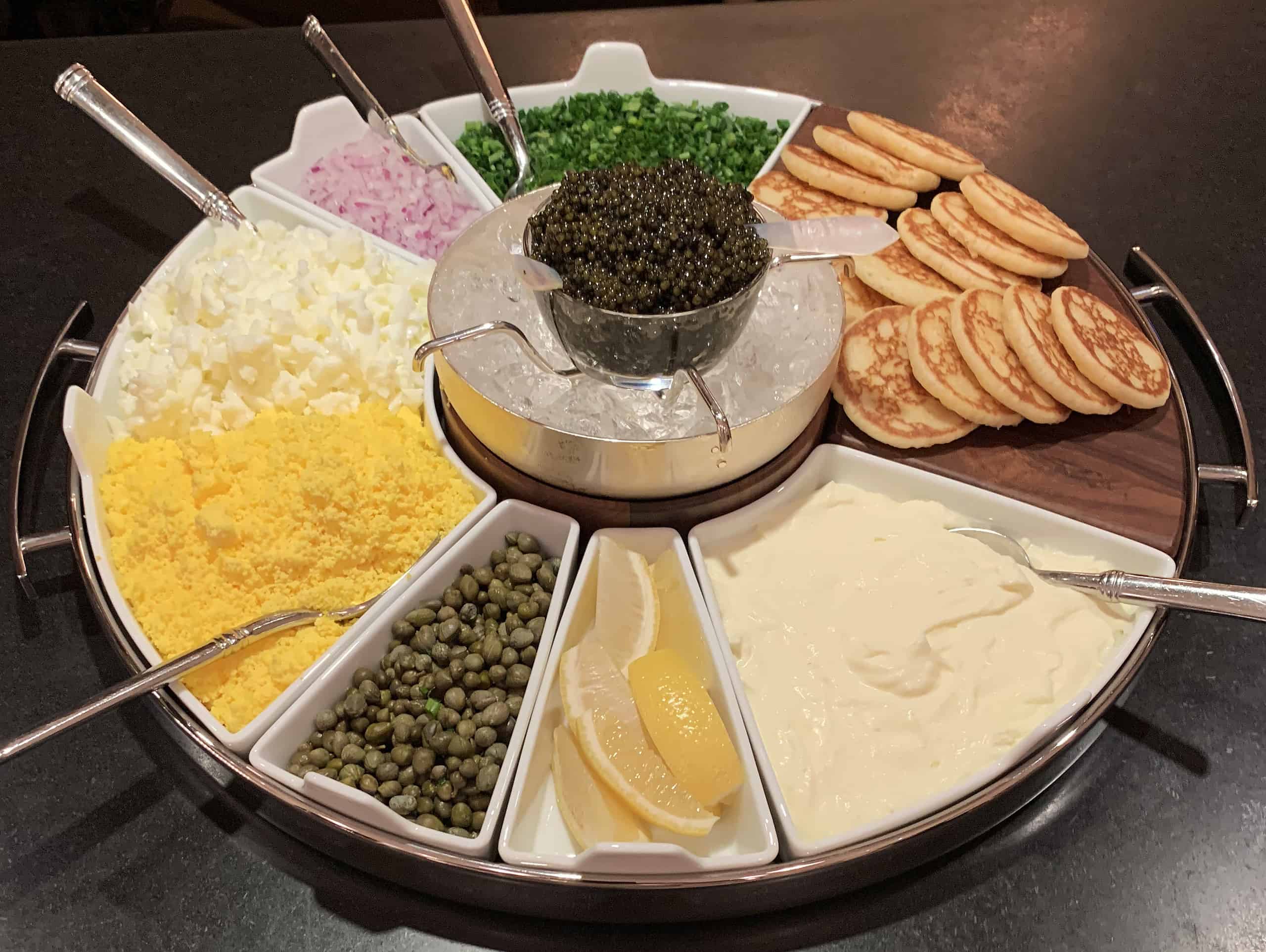 Caviar platter with silver dollar pancakes, chives, chopped red onion, minced cooked egg yolk, lemon, and capers